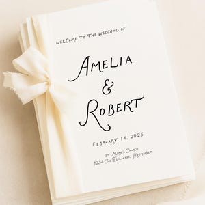 Könnte beinhalten: Ein Stapel weißer Hochzeitskarten, gebunden mit einem cremefarbenen Band. Die Karten zeigen die Namen "Amelia & Robert" in Schreibschrift, das Hochzeitsdatum und den Ort: 14. Februar 2025, St. Mary's Church, 1234 The Esplanade, Haymarket.