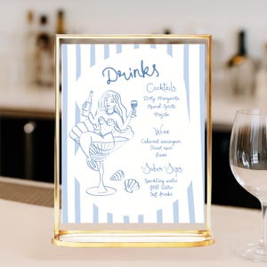 May include: A gold-framed sign with a blue and white striped background. The sign features a mermaid illustration and a list of drinks, including cocktails, wine, and sober sips. The text on the sign reads "Drinks" and lists various beverage options.