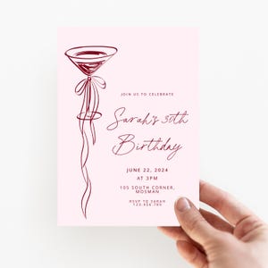 May include: A pink invitation card with a burgundy cocktail glass illustration. The text reads "Sarah's 30th Birthday" with the date, time, and location details. The card is held by a hand.