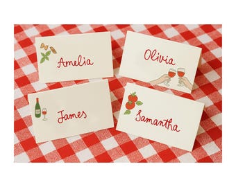 May include: Four cream-colored place cards with names in red script: Amelia, Olivia, James, and Samantha. Each card has a small illustration: pasta, wine glasses, a wine bottle, and tomatoes. The cards are on a red and white checkered tablecloth.