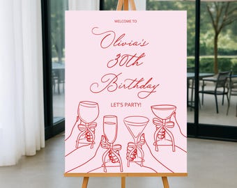 May include: A pink sign on a wooden easel with red text that reads "Welcome to Olivia's 30th Birthday, Let's Party!" The sign features line drawings of hands holding glasses.