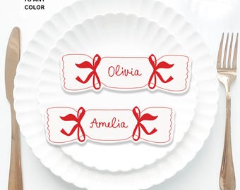 May include: Two white Christmas cracker place cards with red ribbon bows and the names Olivia and Amelia written in red. The cards are on a white plate with a scalloped edge. The maximum size of the cards is 6 inches by 2 inches.