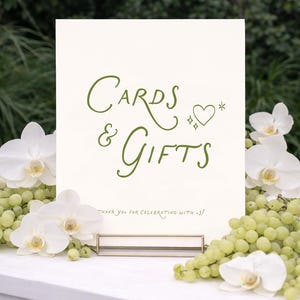 May include: A cream-colored sign with the words "Cards & Gifts" in green script, adorned with a heart and star. The sign is held in a gold-colored metal holder and surrounded by white orchids and green grapes. The text "Thank you for celebrating with us!" is at the bottom.