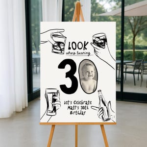 May include: A cream-coloured sign on a wooden easel with black line art of hands holding drinks. The sign reads "LOOK who's turning 30" with a photo of a baby. Text at the bottom says "Let's Celebrate Matt's 30th Birthday."