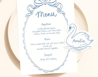 May include: A menu card with a blue bow design at the top and a scalloped oval frame. The menu includes appetizers, main courses, and dessert options. A swan-shaped name card with the name "Amelia" is on the right.