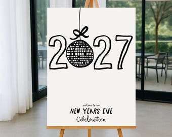 May include: A white sign on a wooden easel with the numbers "2027" in black, a disco ball, and the text "welcome to our NEW YEARS EVE Celebration". The sign is displayed indoors with a blurred outdoor background.