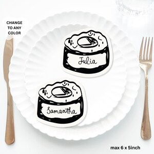 May include: Two black and white sushi-shaped place cards, one labelled "Julia" and the other "Samantha", sit on a white plate. A gold fork and knife are on either side. The text "CHANGE TO ANY COLOR" is in the upper left corner. The dimensions are max 15 x 13 cm.