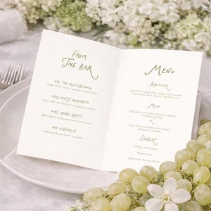 May include: A white menu card with green text, open on a white plate, with grapes and flowers. The menu includes "From The Bar" and "Menu" headings, with drink and food options.