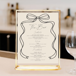 May include: A gold-framed bar menu with a cream-colored background. The menu features black script with cocktail, wine, and mocktail options. A decorative bow and ribbon design tops the menu. A wine glass is visible on the right.