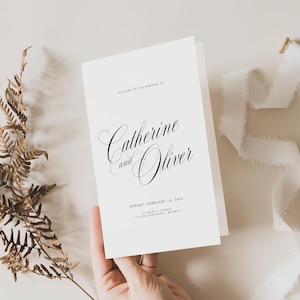 May include: A white folded wedding program card featuring black text that reads Welcome to the wedding of Catherine and Oliver, Sunday, February 14, 2024, St. Mark's Church, Los Feliz, California, with a dried fern and white ribbon.