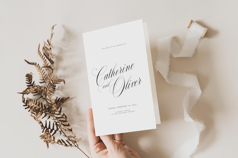 May include: A white folded wedding program card featuring black text that reads Welcome to the wedding of Catherine and Oliver, Sunday, February 14, 2024, St. Mark's Church, Los Feliz, California, with a dried fern and white ribbon.