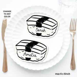 May include: Two sushi-shaped place cards, each with a name written on it, on a white plate. The place cards are black and white, and the plate has a scalloped edge. A knife and fork are on either side of the plate. The text "CHANGE TO ANY COLOR" is in the upper left corner.
