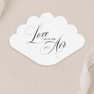 以下が含まれることがあります： エレガントな黒い筆記体で「Love is in the Air」と書かれた、白いスカラップエッジのカード。カードはニュートラルなベージュの背景にあり、隅にはシアーリボンがあります。