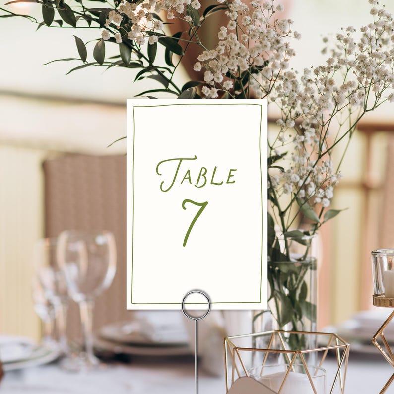May include: A table number card with the word "Table" and the number "7" in green script. The card is held by a silver metal holder. The background includes flowers and glassware.
