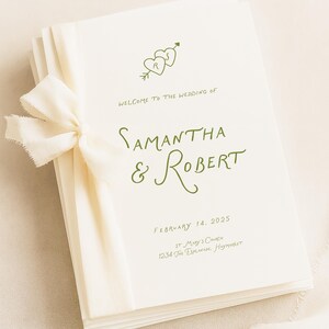 Puede incluir: Una pila de programas de boda de color crema atados con una cinta blanca deshilachada. El texto dice: Welcome to the wedding of Samantha & Robert, February 14, 2025, St Mary's Church, 1234 The Esplanade, Haymarket, con dos corazones y una flecha.