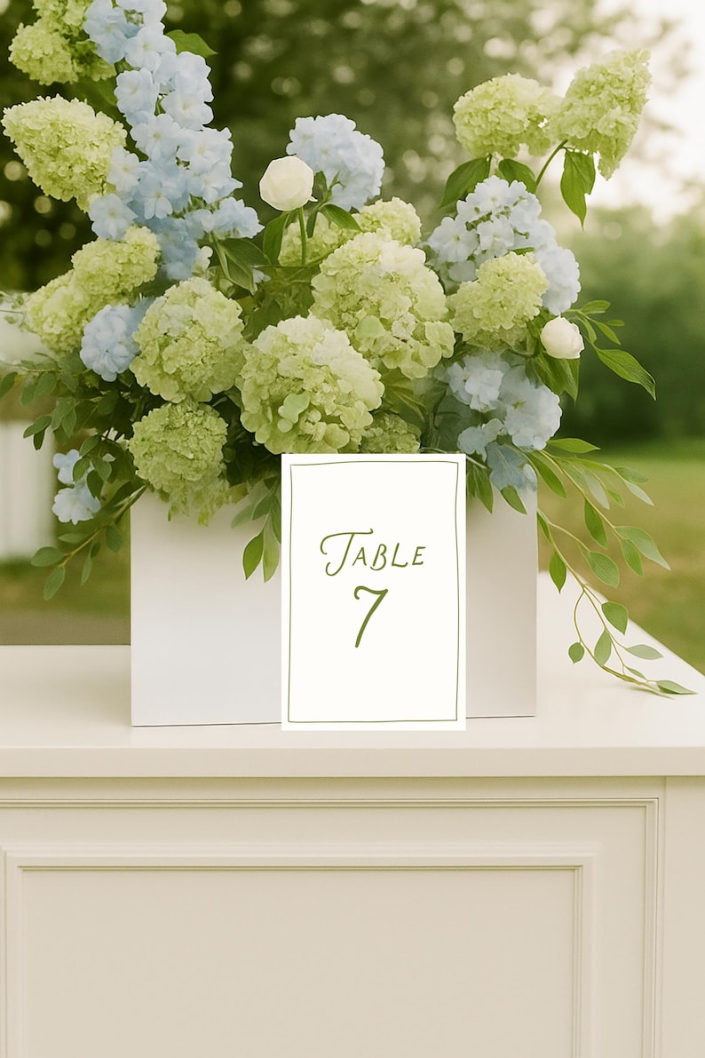 May include: A white rectangular box overflowing with green and blue flowers, with a white table number card that reads "Table 7" in green script. The flowers are a mix of hydrangeas and other greenery, creating a fresh, natural arrangement.