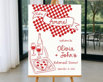 May include: A white sign on a wooden easel with red line art. The sign reads "That's Amore!" and welcomes guests to Olivia + John's Rehearsal Dinner on September 12, 2025. The design includes a checkered pattern, wine, and pizza.