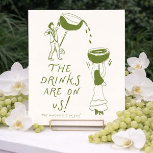May include: A rectangular white sign with olive green line art illustrations of two figures pouring drinks. The text reads THE DRINKS ARE ON US! and below it, in smaller script, THE HANGOVER IS ON YOU! The sign is held by a gold metal stand.