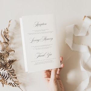 May include: A white folded card featuring black printed text with the headings Reception, Loving Memory, and Thank You. The card is displayed alongside a dried brown fern frond and a piece of white frayed ribbon on a neutral surface.