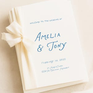 Könnte beinhalten: Ein Stapel cremeweißer Hochzeitskarten, zusammengebunden mit einem ausgefransten cremefarbenen Band. Der Text lautet: Welcome to the wedding of Amelia & Tony, February 14, 2025, St. Mary's Church, 1234 The Esplanade, Haymarket, in blauer Schrift.