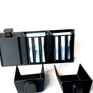 May include: A black 3D printed soap dish with three compartments. The soap dish has a lid with a knob on top. The soap dish is sitting on a white surface.