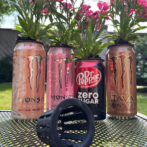 Könnte beinhalten: Drei Aluminiumdosen mit rosa Blumen, die aus ihnen herauswachsen. Die Dosen sind Monster Energy Drinks, Dr Pepper Zero Sugar und Monster Java. Ein schwarzer Plastik-Pflanztopf steht vor den Dosen.
