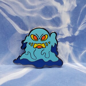 Puede incluir: Un pin de esmalte azul y amarillo de un monstruo de dibujos animados con ojos rojos y una amplia sonrisa. El monstruo tiene la forma de una mancha con un contorno azul oscuro.