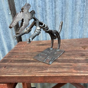 Peut inclure: Une sculpture en métal d'un chien faite à partir d'outils et de quincaillerie recyclés. La sculpture est posée sur une surface en bois et comporte une plaque en métal avec les lettres "CH" dessus.