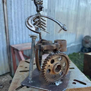 May include: A metal sculpture of a skeleton playing the drums. The skeleton is wearing a hat and has a horseshoe for a ribcage. The drums are made of metal gears and pipes.