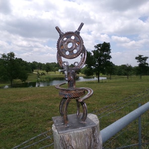 May include: A rusty metal sculpture of a person made from gears, horseshoes, and other metal pieces. The sculpture is mounted on a wooden post and is in front of a green field with a pond in the background.
