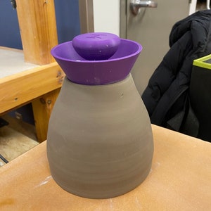 Può includere: Un vaso in ceramica di colore viola con un coperchio viola. Il coperchio ha un piccolo pomo in cima.