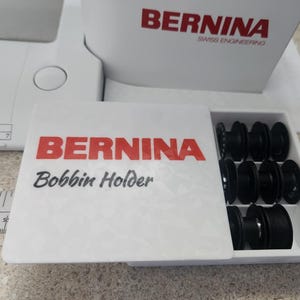 Könnte beinhalten: Ein weißer Bernina-Spulenhalter mit rotem Markennamen. Der Halter ist geöffnet und zeigt mehrere schwarze Spulen. Der Text "Bobbin Holder" ist darunter gedruckt.