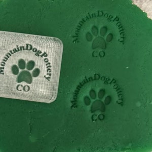 May include: A clear plastic stamp with the text "Mountain Dog Pottery CO" and a paw print design. The stamp is on a green surface.