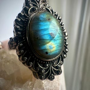 May include: A silver ring featuring an oval-shaped labradorite gemstone with blue and yellow iridescence. The metal band is decorated with intricate scrollwork and bead-like details surrounding the central stone in a symmetrical pattern.