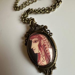 May include: A bronze chain necklace with an oval pendant featuring a vintage illustration of a woman's profile in red and black.