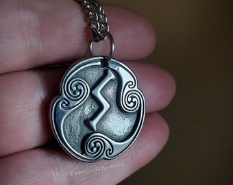 May include: Silver-toned pendant necklace featuring a stylized lightning bolt design within a circular frame. The pendant has intricate scrollwork around the edges and hangs from a delicate chain. The necklace is held in a hand against a blurred background.