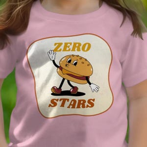 May include: Pink t-shirt featuring a cartoon hamburger character with the words "ZERO STARS" in a retro font. The burger has a smiling face, arms, and legs, waving with a white-gloved hand. The design is set within a cream-colored, rounded rectangle.