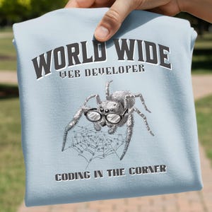 May include: Light blue t-shirt with the text "WORLD WIDE WEB DEVELOPER" and "CODING IN THE CORNER". The shirt features a detailed illustration of a spider wearing glasses and sitting on a web. The design is in black and white.