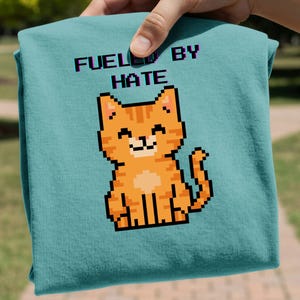 May include: A teal t-shirt featuring a pixelated orange cat with the text "FUELED BY HATE" in a retro font. The cat is smiling. The shirt is held up by a hand.