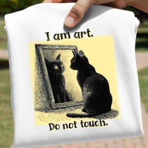 May include: White t-shirt featuring a black cat looking in a mirror at its reflection. The mirror is in a decorative frame, and the image has the text "I am art. Do not touch."