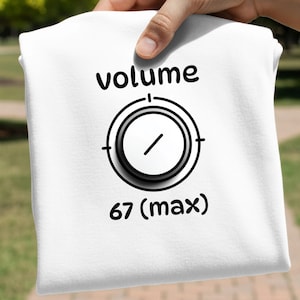 May include: White t-shirt with a black graphic of a volume knob and the word "volume" above it. Below the knob is the text "67 (max)". The shirt is held by a hand.