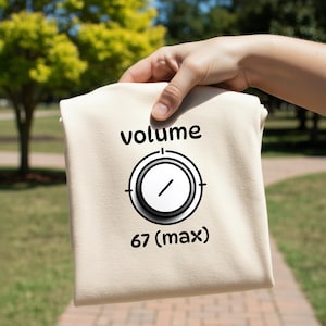 May include: A beige t-shirt with the word "volume" above a graphic of a volume knob. Below the knob is the text "67 (max)". The t-shirt is held up by a hand against a blurred background of green trees and a brick walkway.