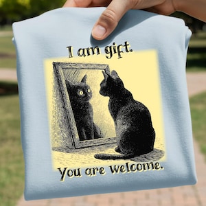 May include: Light blue t-shirt featuring a black cat looking in a mirror at its reflection. The mirror image and the cat are black. The text "I am gift. You are welcome." is printed on the shirt.