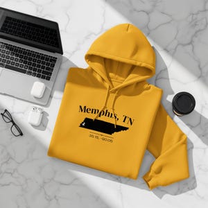May include: Mustard yellow hoodie with the text "Memphis, TN" and a black graphic of the city's outline. The coordinates 35.15, -90.05 are printed below the text. A laptop, glasses, and a coffee cup are also in the image.