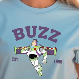 A light blue t-shirt with a graphic of Buzz Lightyear, the main character from the Toy Story franchise, and the text "BUZZ EST 1995" printed on it.