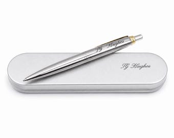 May include: A silver-toned ballpoint pen with a gold-colored clip and the engraved name "JG Hughes" resting on a matching silver-toned metal rectangular case, which also features the engraved name "JG Hughes" on its top surface.