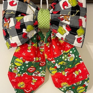 May include: A large fabric bow featuring a black and white buffalo plaid pattern with Grinch and Max illustrations on the top loops. The center is a light green fabric with white polka dots. The two long tails feature red and green panels.