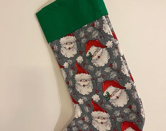 Puede incluir: Un calcetín navideño con fondo gris y un patrón repetitivo de caras de Papá Noel con gorros rojos, decorado con pequeñas bayas rojas y hojas de acebo grises. El calcetín presenta un puño verde sólido y una trabilla de tela gris a juego.