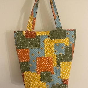 May include: A quilted tote bag featuring a patchwork design of various fabric squares in shades of olive green, mustard yellow, orange, and light blue. The fabric patterns include small white dots, yellow sunflowers, and checkered prints with visible stitching.