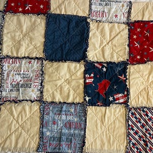 Puede incluir: Un edredón de patchwork con temática patriótica. El edredón presenta cuadrados de tela crema, roja y azul marino. Algunos cuadrados tienen patrones de estrellas y rayas, otros tienen texto relacionado con la Independencia Americana.